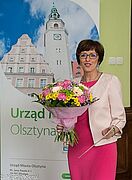 Jolanta Okuniewska z bukietem kwiatów na tle bannera miasta Olsztyna
